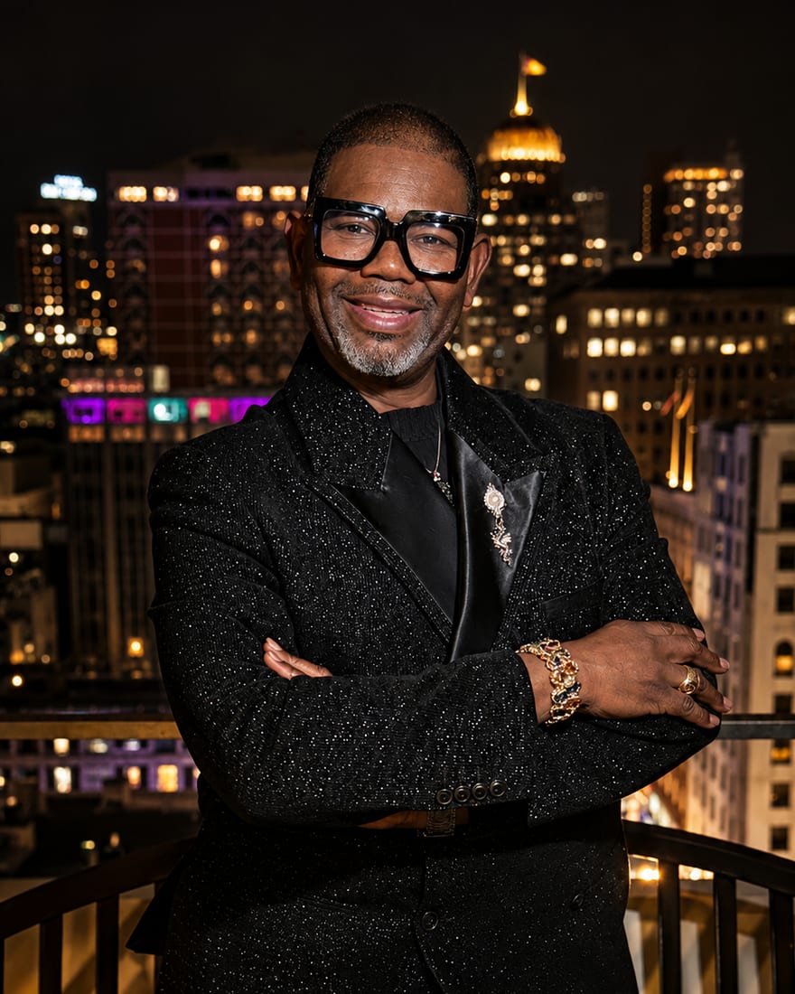 John Weber in a black sparkle-flecked jacket, arms crossed, on a rooftop balcony at night with the lit San Francisco skyline behind him