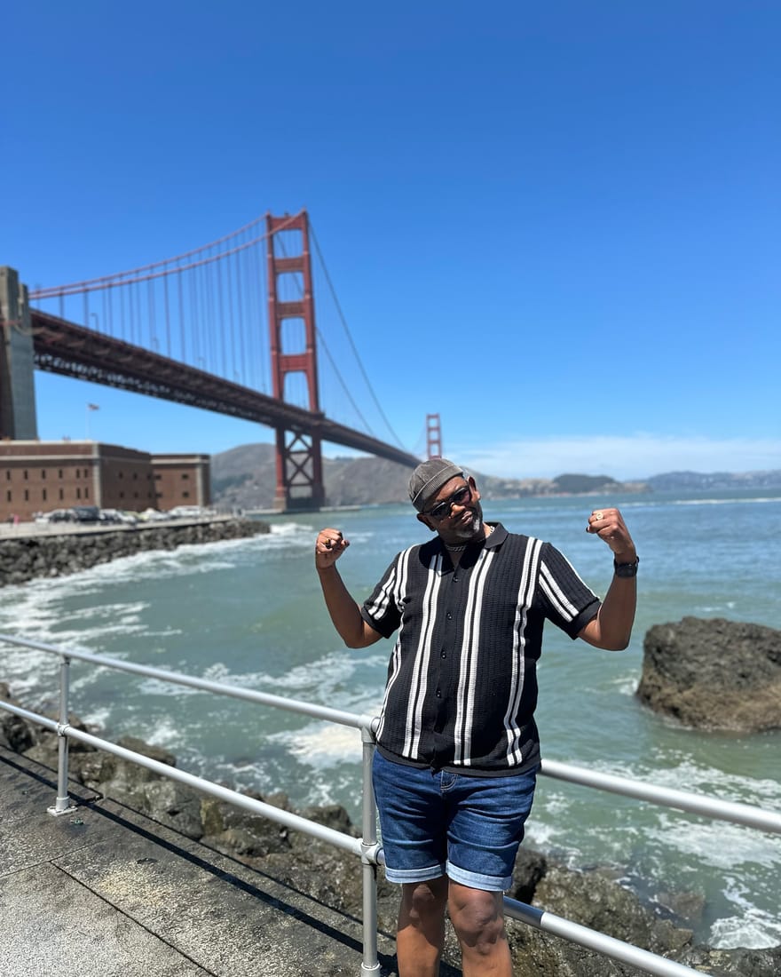 John Weber flexing playfully on the waterfront at Fort Point in San Francisco, with the Golden Gate Bridge and the bay behind him