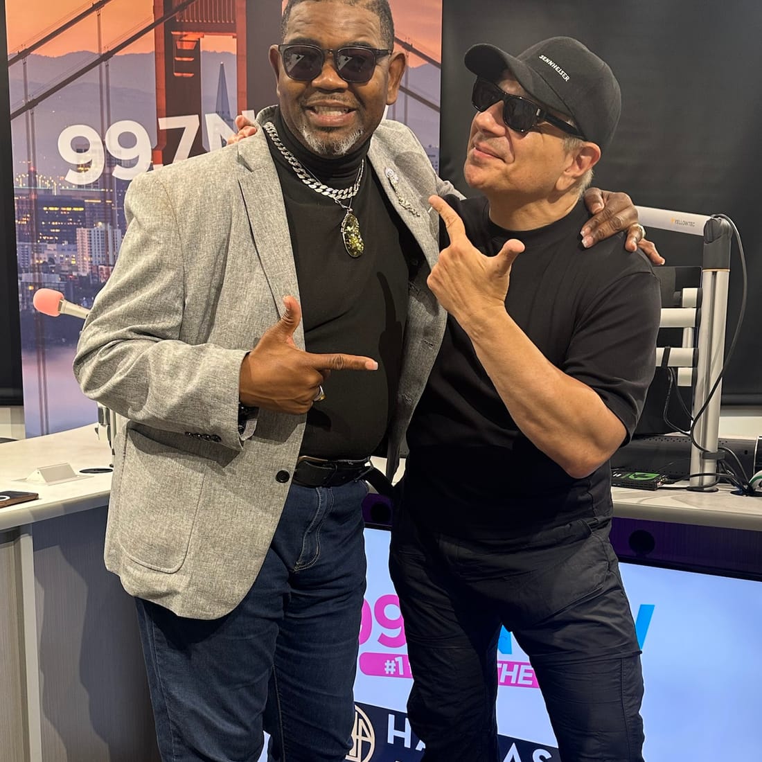 John Weber in a gray blazer and black turtleneck with a radio host in the 99.7 NOW studio in San Francisco, a Golden Gate Bridge backdrop behind them